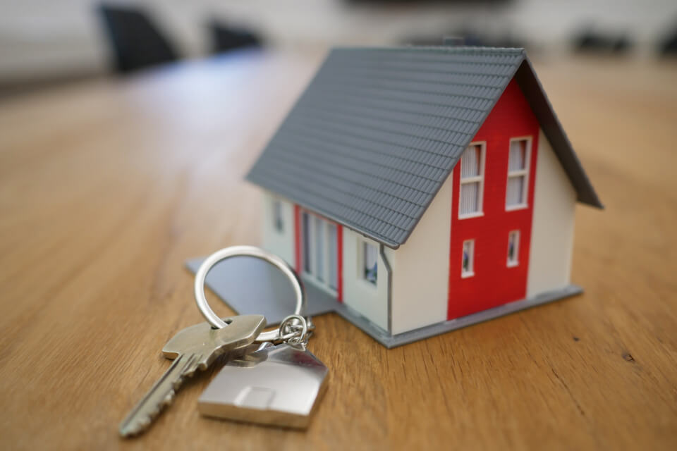 House key with white tag resting on wooden surface next to miniature model house with red walls and gray roof