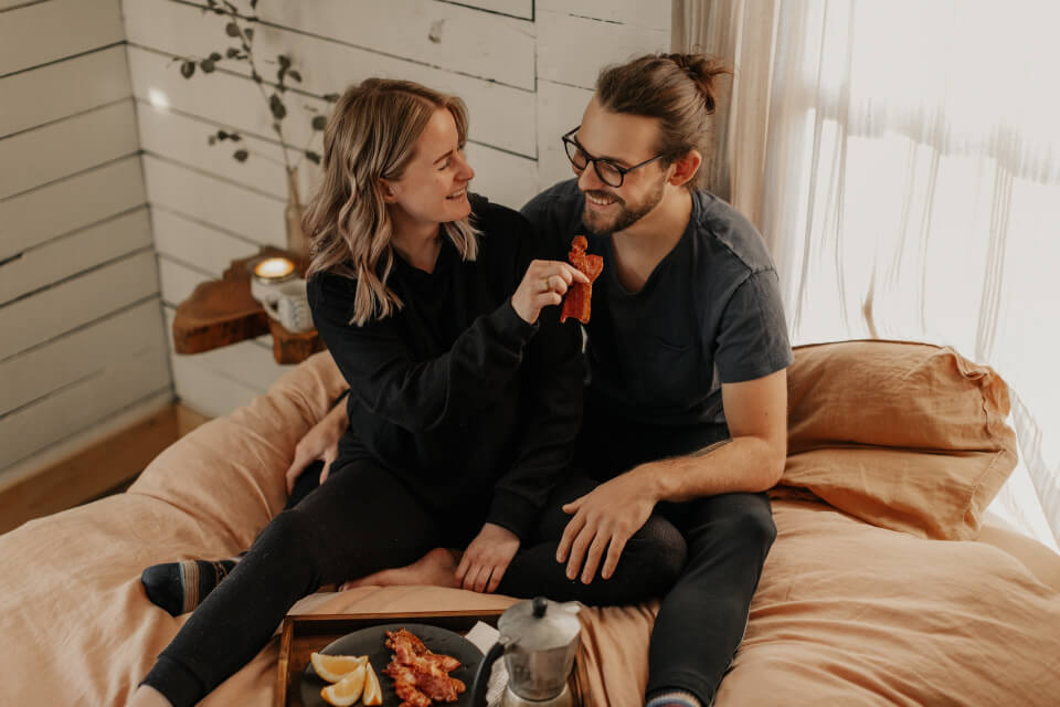 Couple smiling and sharing food together on a bed in a modern bedroom with white shiplap walls and warm lighting