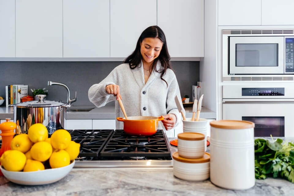 Woman cooking in a modern kitchen while stirring a red pot on a gas stove, with fresh lemons and kitchen accessories visible on the countertop