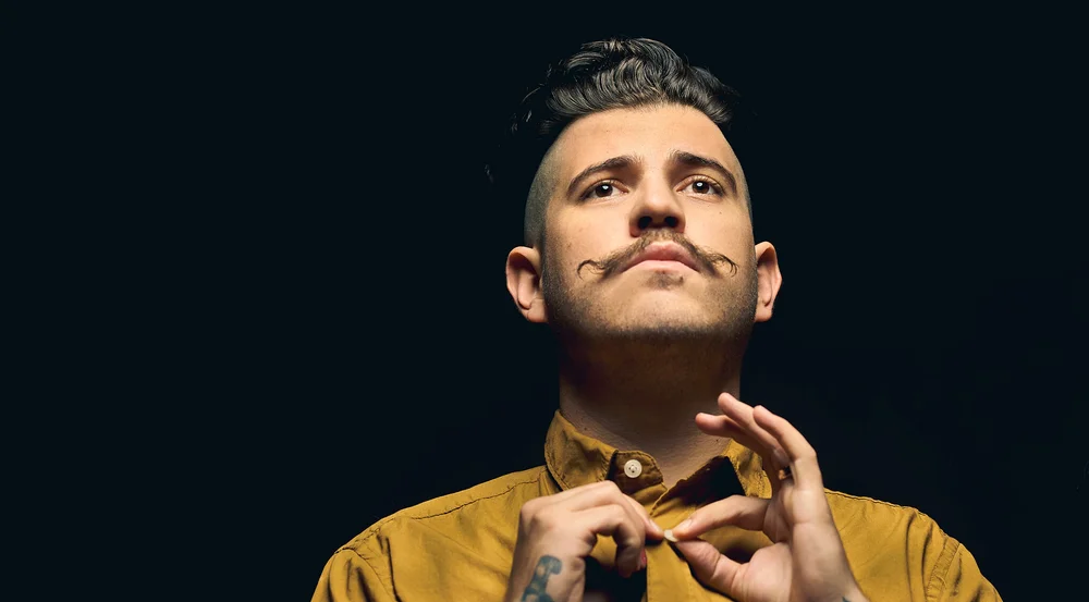 Handsome man with styled mustache wearing a golden yellow shirt against a dark background, photographed by Erik Pinkerton Photography