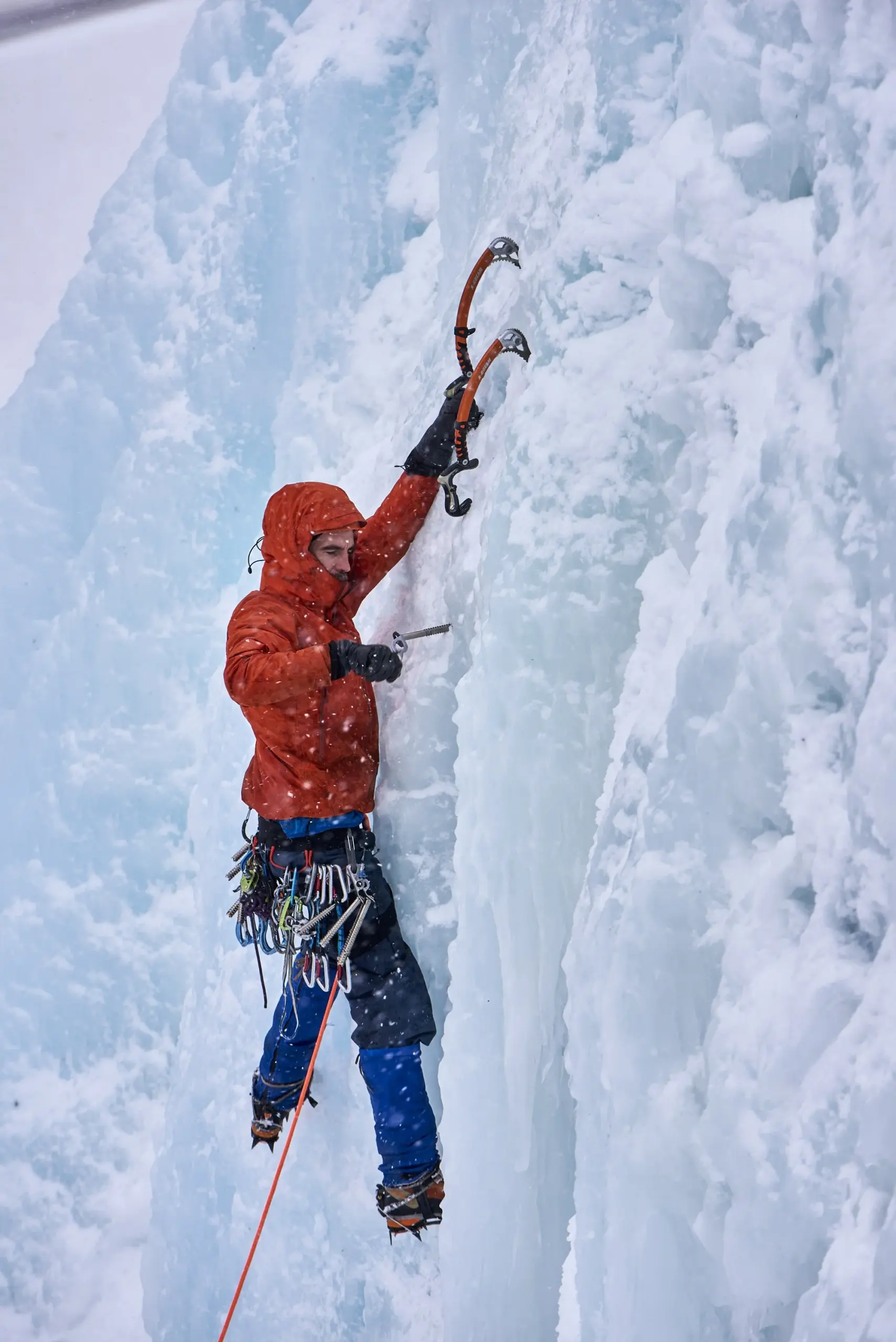 Ice climber in red jacket and blue pants scaling a frozen waterfall with ice axes and climbing rope, surrounded by white and blue ice formations