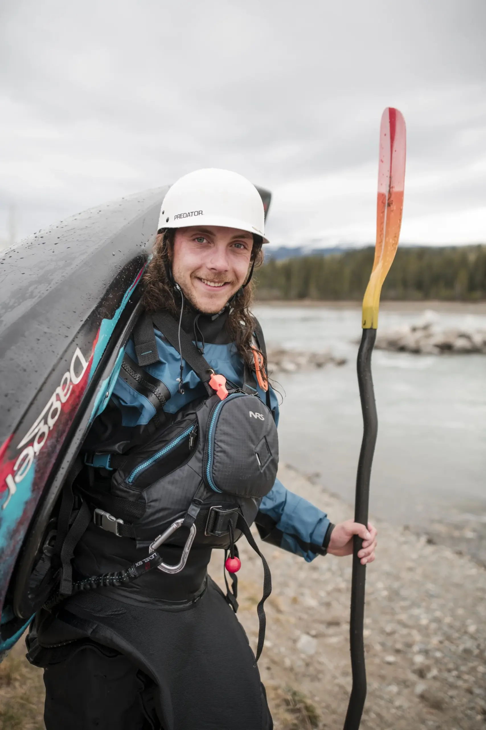 Smiling kayaker wearing a white Sedator helmet and blue life jacket holds a colorful paddle while carrying a kayak at a riverbank with forested mountains in the background