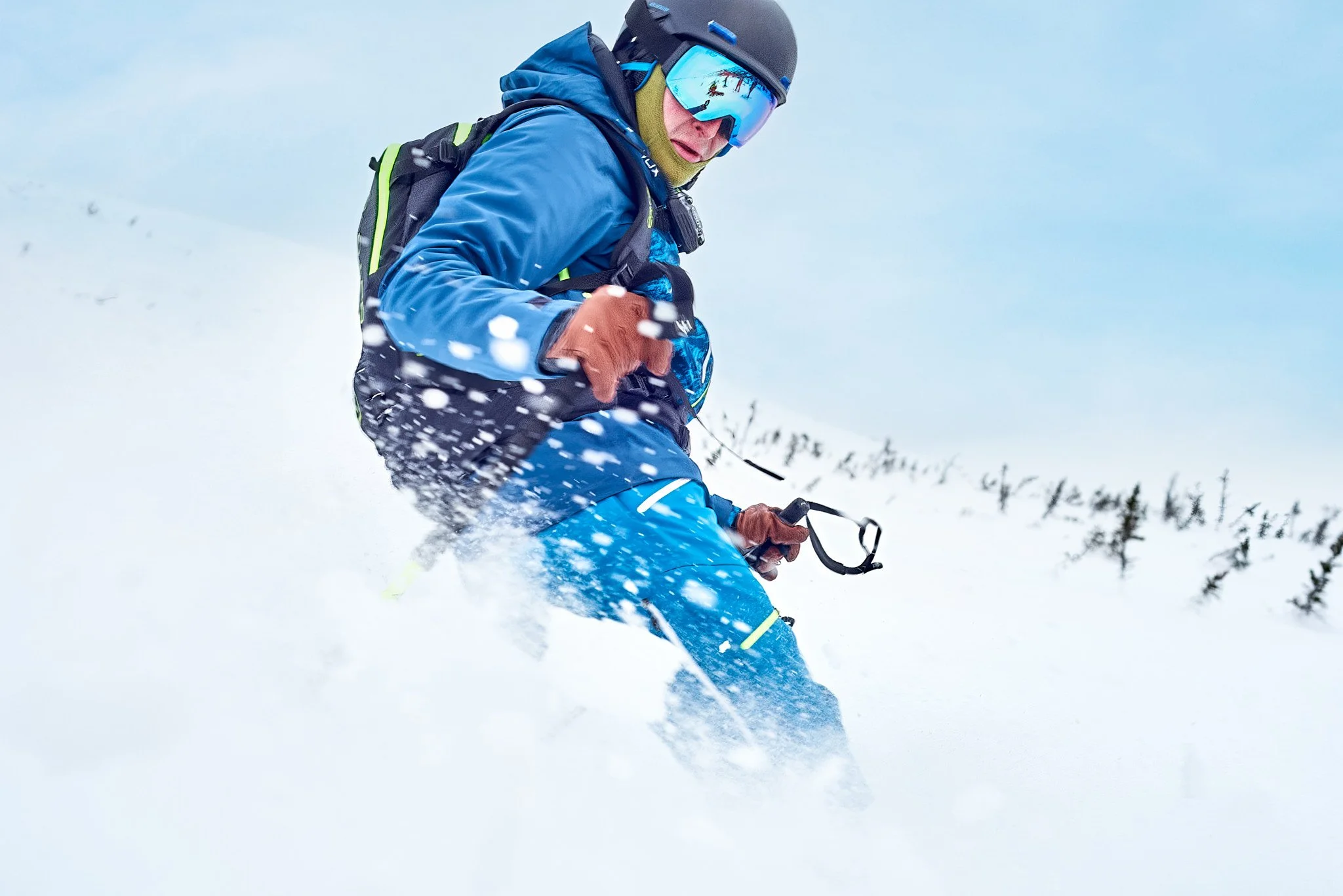 A snowboarder in blue jacket and helmet riding down a snowy mountain slope with powder spraying, forest visible in the background