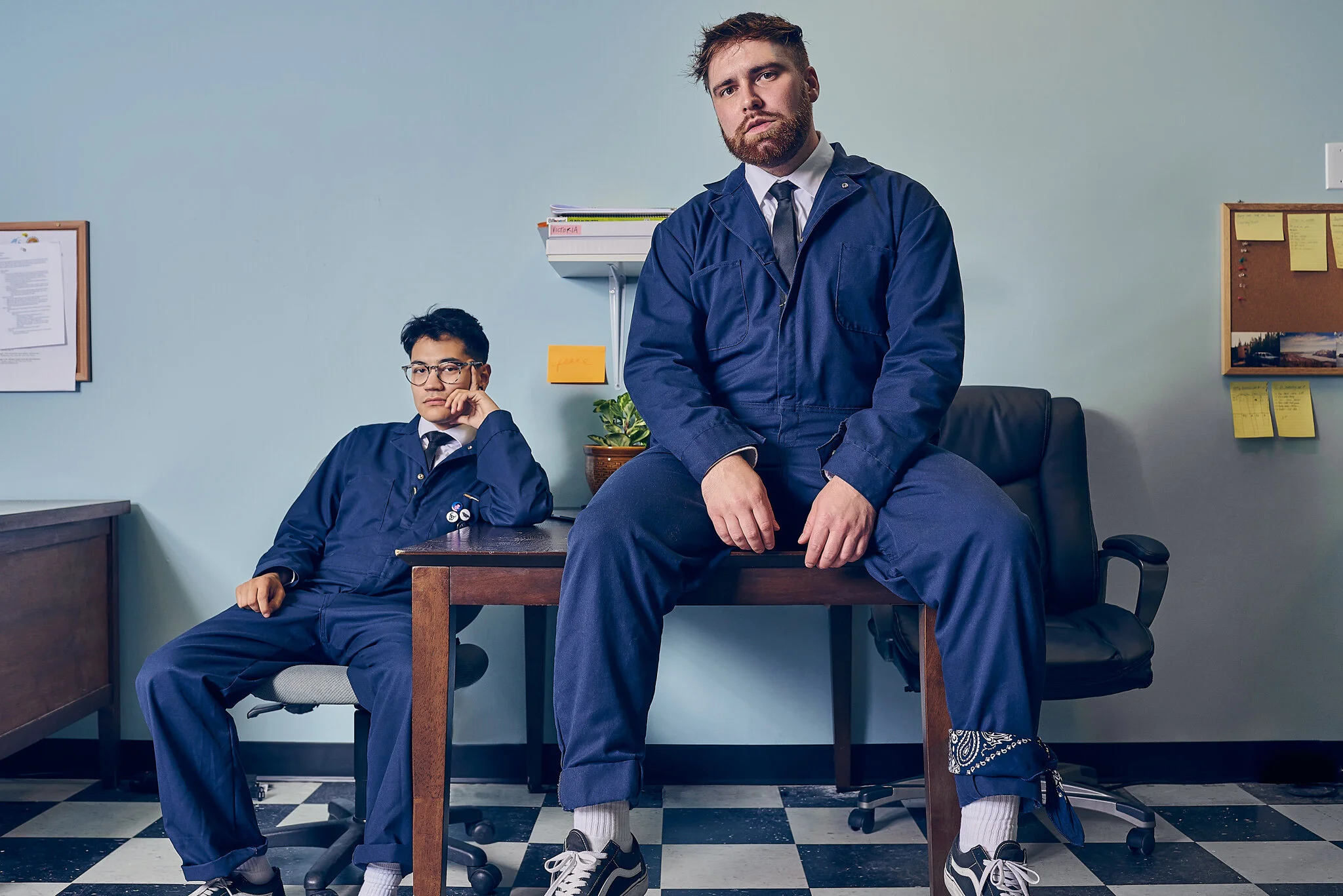 Two men in blue uniforms sitting on a desk in an office setting with bulletin boards and sticky notes on the walls