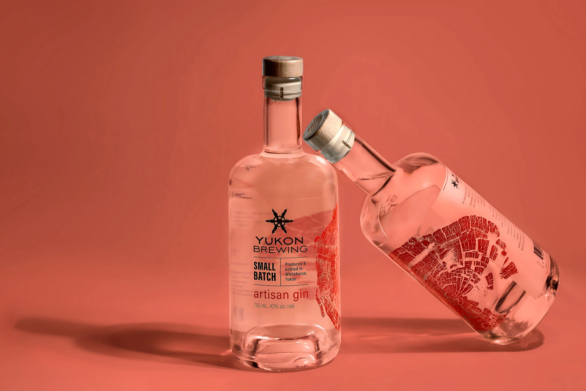 Two clear glass bottles of Yukon Brewing Small Batch artisan gin with cork stoppers displayed against a coral-pink background, one bottle upright and one tilted